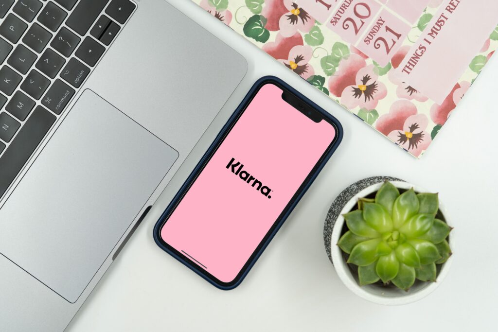 Flat lay photo of a mobile phone with the Klarna App open on screen with a laptop and calendar nearby, representing the idea of using Buy Now Pay Later options when shopping online.