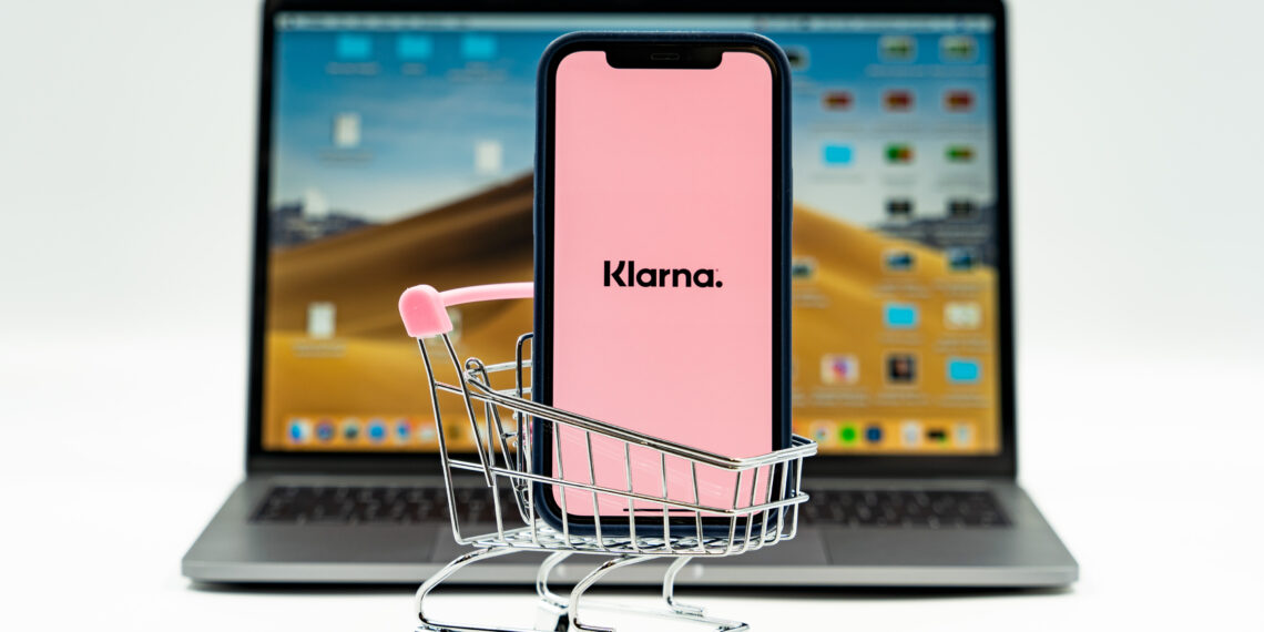 Klarna App open on a mobile phone placed inside a small shopping trolley, with the Klarna website open on a laptop in the background.