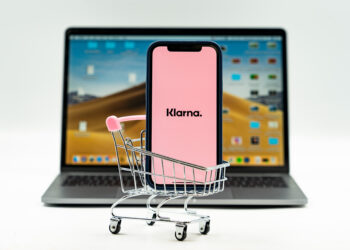 Klarna App open on a mobile phone placed inside a small shopping trolley, with the Klarna website open on a laptop in the background.