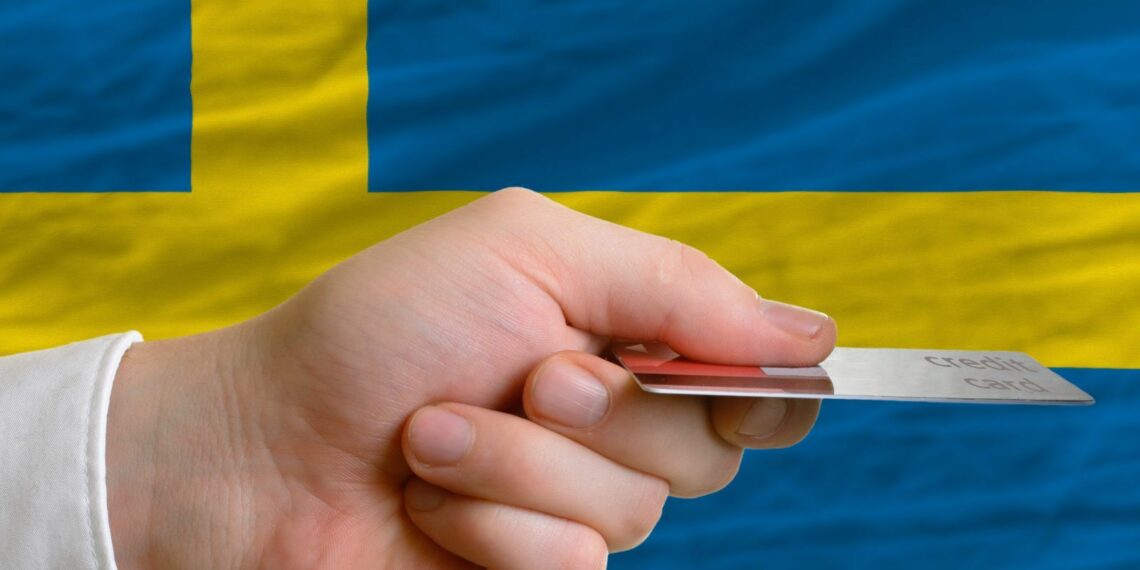 A hand holding a credit card and Sweden's flag is in the background.